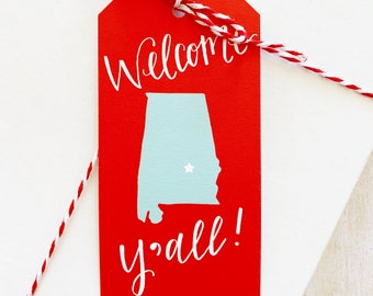 May include: Red gift tag with white text that says "Welcome y'all!" and a blue outline of the state of Alabama with a white star in the center.