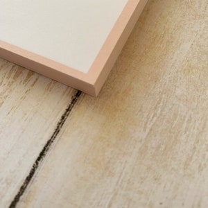May include: A close-up of the corner of a white card with a light pink border. The card is lying on a light brown wooden surface.