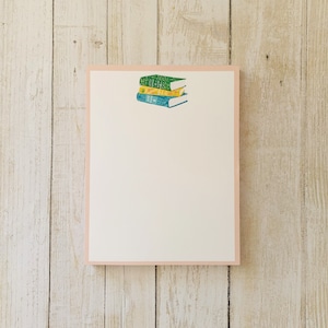 May include: A white notepad with a pink border and a stack of three books with green, yellow, and blue covers.