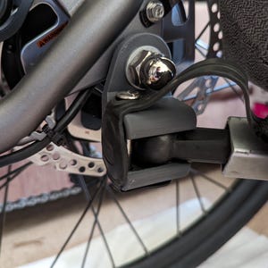 May include: A black metal bike trailer hitch with a silver metal bracket and a black strap. The hitch is attached to a bicycle.