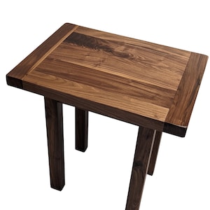 May include: A small, rectangular wooden table with a rich, dark brown finish. The tabletop has a framed border and is supported by four sturdy legs. The table is approximately 24 inches long, 18 inches wide, and 20 inches tall.
