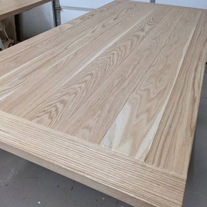 May include: A light wood table top with a natural finish. The table top is made of several planks of wood that are joined together. The table top has a simple, rectangular shape.