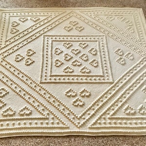 Puede incluir: Una manta de ganchillo de color crema con un patrón geométrico de corazones y círculos. La manta tiene una superficie texturizada y en relieve.