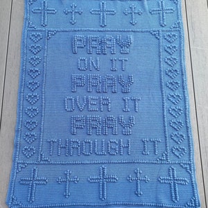 May include: A blue crocheted blanket with a white cross pattern border. The centre of the blanket features the text "Pray On It Pray Over It Pray Through It".