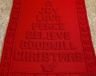 May include: Red crocheted blanket with a Christmas tree design. The tree is made of raised stitches and features the words "Joy", "Love", "Peace", "Believe", "Goodwill", and "Christmas".