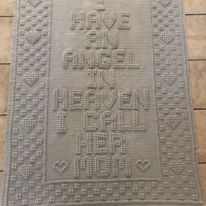 Könnte beinhalten: Eine graue gehäkelte Decke mit den Worten "Ich habe einen Engel im Himmel, von dem ich dir nie erzählt habe" in erhabenen Buchstaben.