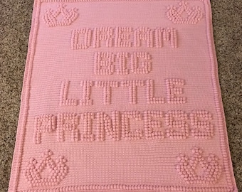 May include: A pink crocheted baby blanket with the words "DREAM BIG LITTLE PRINCESS" in raised letters. The blanket features four crown designs in the corners. The blanket is made of soft yarn and is perfect for a baby girl.