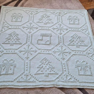 May include: A light blue crocheted blanket with a repeating pattern of Christmas trees, houses, and stockings. The blanket is made with a textured stitch that gives it a raised, puffy look.