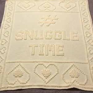 May include: A white crocheted blanket with the text "#SNUGGLE TIME" in raised lettering. The blanket has a border of hearts and diamonds.
