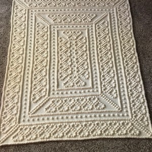 Könnte beinhalten: Cremefarbene, quadratische Häkeldecke mit detailliertem geometrischem Muster. Das Design weist erhabene Noppen und strukturierte Stiche auf, wodurch eine taktile Oberfläche entsteht. Die Decke hat einen Rand mit sich wiederholendem Muster und ein zentrales quadratisches Design.