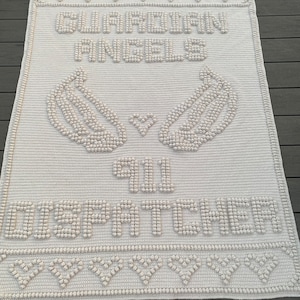 Pode incluir: Uma manta de crochê branca com as palavras "Guardian Angels" e "Still Our Hearts" em letras em relevo.