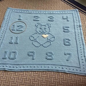 Può includere: Una copertina per neonati in crochet blu con un orsacchiotto bianco al centro e i numeri da 1 a 12 intorno al bordo.