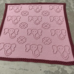 May include: A handmade pink crochet blanket featuring an elephant and flower pattern. The blanket has a textured design with a dark red border. This soft, cozy blanket is perfect for a baby or child.