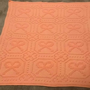 May include: A peach-coloured crocheted blanket with a repeating pattern of hearts. The blanket is made with a textured yarn that gives it a raised, puffy look.