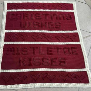 May include: A handmade, red and white crocheted blanket with the words "CHRISTMAS WISHES" and "MISTLETOE KISSES" in raised letters. The blanket features heart-shaped patterns and a white border, perfect for holiday decor.