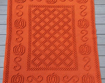 May include: An orange crochet blanket with a pumpkin theme. The blanket features a central diamond pattern, a border of pumpkins, and decorative swirls. The blanket is made of a soft, textured yarn, perfect for fall decor.