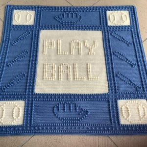 以下が含まれることがあります： 白い糸で「PLAY BALL」と書いてある白い中央パネルが付いた青い編み物の毛布。毛布は白い野球ボールの縁取りがされている。