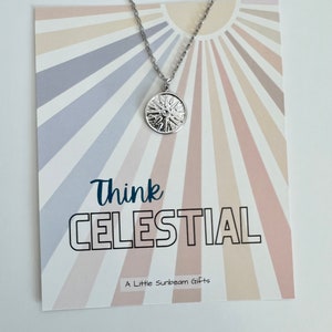 May include: A silver-colored necklace with a circular pendant featuring a sunburst design. The pendant hangs from a delicate chain. The necklace is displayed on a card with the words "Think CELESTIAL" and "A Little Sunbeam Gifts" printed on it.