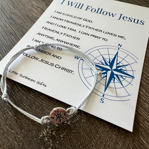 May include: White string bracelet with a compass charm. The bracelet is tied in a knot and has the words "I Will Follow Jesus" and "Follow Jesus Christ" on a white card with a compass design.