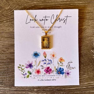 May include: A gold-colored necklace with a rectangular pendant featuring a sunburst design, displayed on a card with floral illustrations. The card reads "Look unto Christ" and includes a Bible verse. The brand name "A Little Sunbeam Gifts" is also visible.