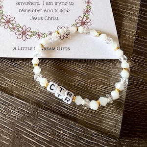May include: A white beaded bracelet with gold accents and clear crystal beads. The bracelet spells out "CTR" with white letter beads. The background includes a card with text and floral designs.