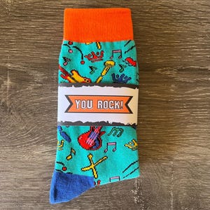 May include: A pair of socks with a teal background and musical instrument graphics, including guitars and microphones. The cuff is orange, the toe is blue, and a white band across the middle reads "YOU ROCK!"