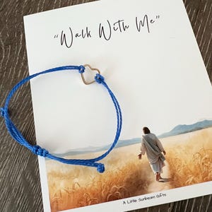 May include: A blue cord bracelet with a silver heart-shaped charm rests on a white card. The card features the text "Walk With Me" and an illustration of a figure walking through a field. The words "A Little Sunbeam Gifts" are at the bottom.