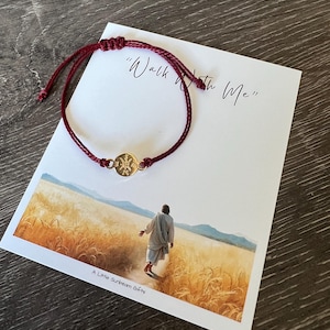 May include: A burgundy cord bracelet with a gold-colored circular charm. The bracelet is displayed on a white card with the text "Walk With Me" and an image of a person walking in a field. The card also has the text "A Little Sunbeam Gifts".