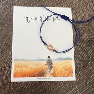 May include: A navy blue adjustable bracelet with a gold sun charm, displayed on a white card. The card features the text "Walk With Me" and an illustration of a figure walking through a wheat field. The text "A Little Sunbeam Gifts" is at the bottom.