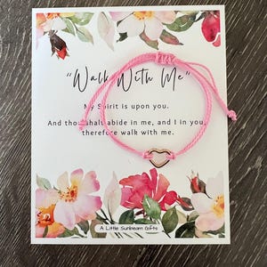 May include: A pink cord bracelet with a gold heart charm is displayed on a card with floral illustrations and the text "Walk With Me". The card also includes the text "My Spirit is upon you" and "A Little Sunbeam Gifts".