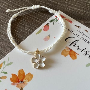 May include: White braided bracelet with a white flower charm. The bracelet is on a white card with the text "I can do all things through Christ who strengthens me" and floral illustrations.