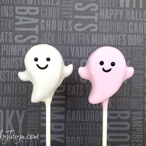 May include: Two ghost-shaped treats on sticks, one white and one pink, with black smiling faces. The treats are set against a gray background with Halloween-themed words. The text "TreatsbyTaryn.com" is at the bottom.