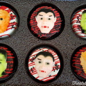 May include: A box of six Halloween-themed decorated cookies. Each cookie features a different monster face, including Frankenstein's monster, a vampire, and a werewolf. The cookies are on a white base with red and black accents.