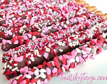 May include: Chocolate-covered pretzel rods dipped in chocolate and decorated with pink, red, and white sprinkles and heart-shaped sprinkles. The pretzels are arranged in rows on a white surface. TreatsbyTaryn.com is visible in the bottom right corner of the image.
