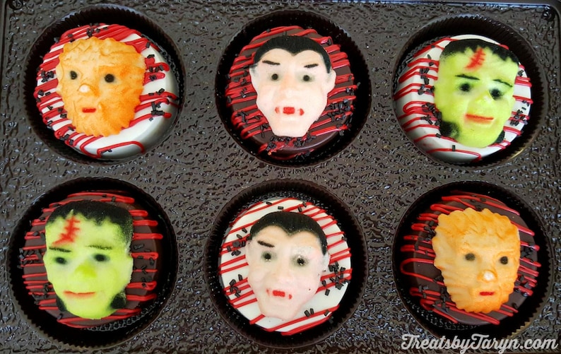 May include: A box of six Halloween-themed decorated cookies. Each cookie features a different monster face, including Frankenstein's monster, a vampire, and a werewolf. The cookies are on a white base with red and black accents.