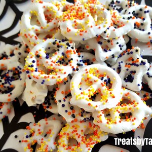 May include: White chocolate-covered pretzels with colorful sprinkles, arranged on a black and white spiderweb design. The pretzels are decorated with orange, yellow, and blue sprinkles. The image includes the text "Treats by Taryn."