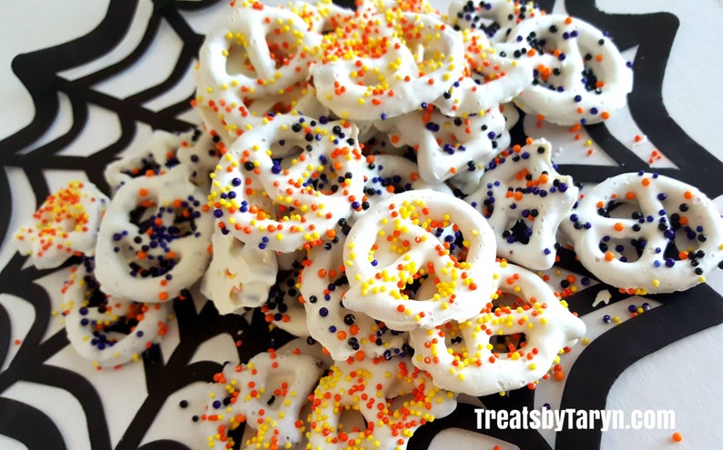May include: White chocolate-covered pretzels with colorful sprinkles, arranged on a black and white spiderweb design. The pretzels are decorated with orange, yellow, and blue sprinkles. The image includes the text "Treats by Taryn."