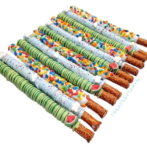 May include: A group of pretzel rods dipped in white chocolate and decorated with green, blue, and white stripes, colorful sprinkles, and watermelon-shaped candies. The pretzel rods are arranged in a diagonal line, with the text "Treats by Taryn" visible in the lower right corner.
