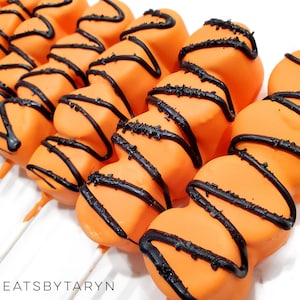 May include: Orange and black marshmallow pops decorated with black icing and sprinkles. The pops are on white sticks and arranged in a row. The text "TREATSBYTARYN" is visible in the bottom left corner of the image.