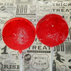 May include: Two round, bright red lollipops on white sticks. The lollipops have a clear, glossy appearance with white flecks. The background features a Halloween-themed pattern with text and illustrations.