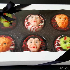 May include: A white box with a clear lid, tied with a black ribbon, containing six Halloween-themed treats. Each treat features a different monster face, including Frankenstein, a werewolf, and a vampire. The treats are decorated with red, white, and black icing.