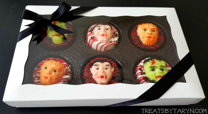 May include: A white box with a clear lid, tied with a black ribbon, containing six Halloween-themed treats. Each treat features a different monster face, including Frankenstein, a werewolf, and a vampire. The treats are decorated with red, white, and black icing.