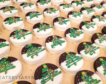 May include: Christmas-themed Oreo cookies, half-dipped in white chocolate, decorated with green icing Christmas trees and colorful sprinkles. The cookies are arranged on a light brown surface.