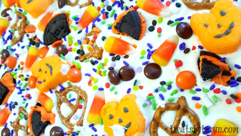 Puede incluir: Primer plano de un postre tem&aacute;tico de Halloween con glaseado blanco, decorado con caramelos de ma&iacute;z, malvaviscos naranjas con forma de calabaza, caramelos de chocolate, pretzels y chispas de colores. El texto "treatsbylaryn.com" est&aacute; visible.