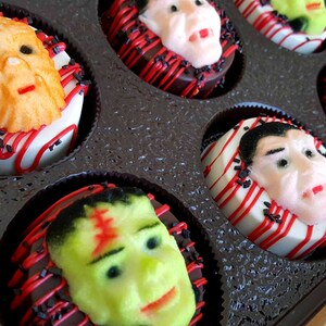 May include: A box of Halloween-themed donuts. Each donut is decorated with a different monster face, including Frankenstein, Dracula, and a werewolf. The donuts are white with red and black accents.