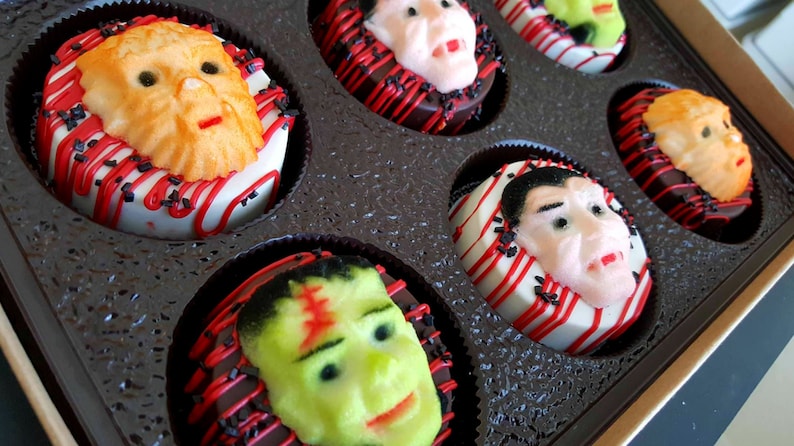 May include: A box of Halloween-themed donuts. Each donut is decorated with a different monster face, including Frankenstein, Dracula, and a werewolf. The donuts are white with red and black accents.