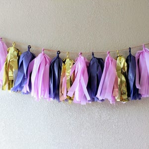 Puede incluir: Una guirnalda de borlas de papel de seda de color morado, rosa y dorado. La guirnalda está enhebrada en una cuerda fina y es perfecta para una fiesta o celebración.