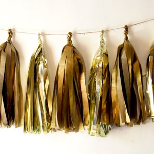 Op de afbeelding: Een slinger van gouden metallic kwasten hangt aan een dunne, natuurlijk gekleurde draad. De kwasten zijn gemaakt van glanzende, gelaagde gouden stroken, wat een feestelijke en decoratieve uitstraling geeft. De kwasten hangen tegen een witte achtergrond.