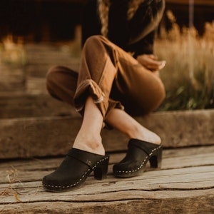 Puede incluir: Zuecos de cuero negro con tacón de madera y una correa en la parte superior. Los zuecos los lleva puestos una persona sentada en unos escalones de madera.