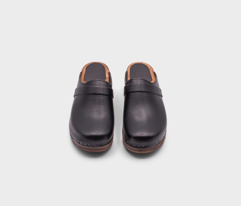 Malmö-style clogs designed by Sandgrens in classic black leather. Featuring a smooth, slip-on design with a supportive strap for stability. The sturdy wooden sole and hand-stapled construction ensure durability and all-day comfort, making them a stylish and functional footwear choice.